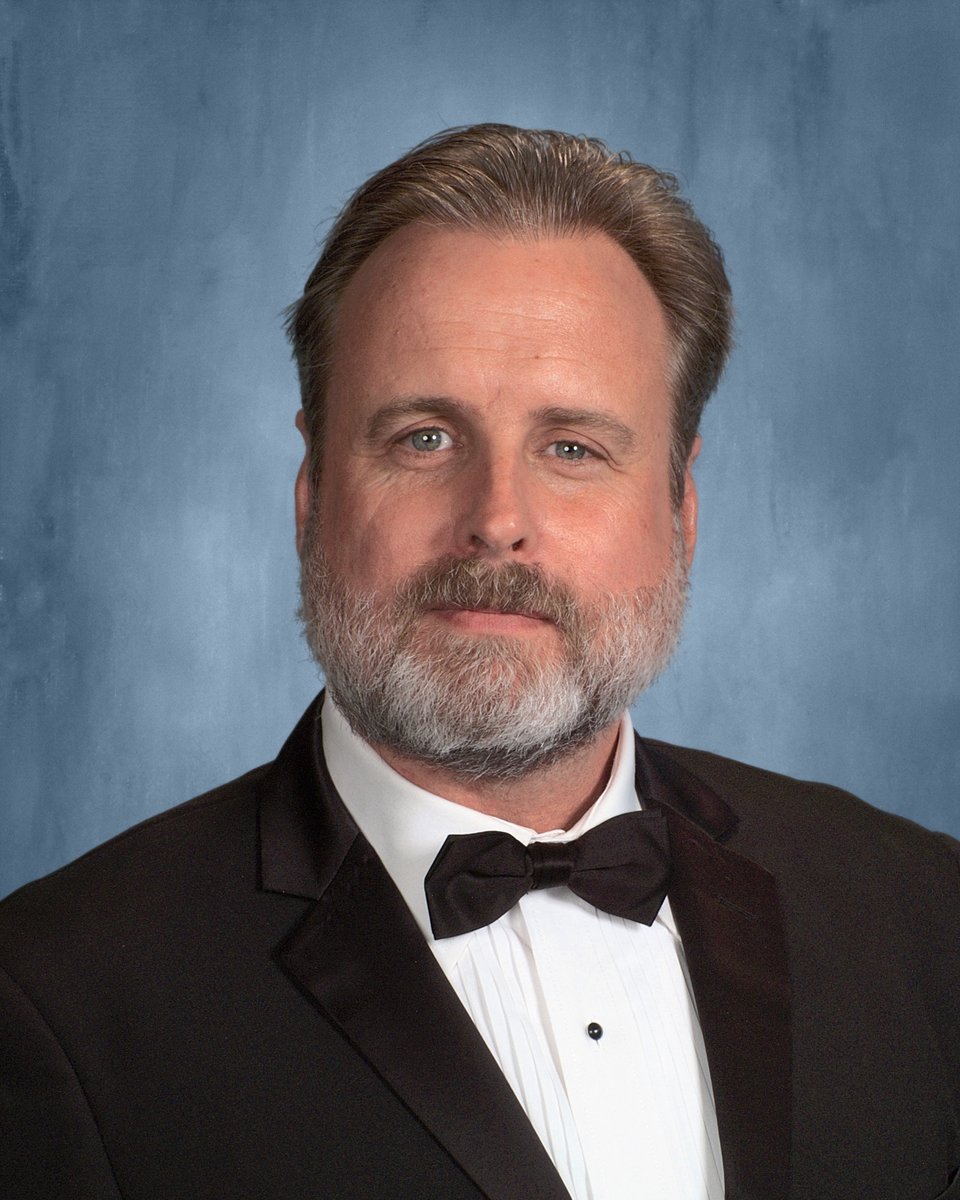 Steve Willis, in a black tuxedo and bow tie, poses for a formal portrait against a blue studio backdrop