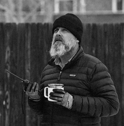 Steve Willis, in a knit cap and down jacket, looks upward while holding a handheld radio and a mug of coffee