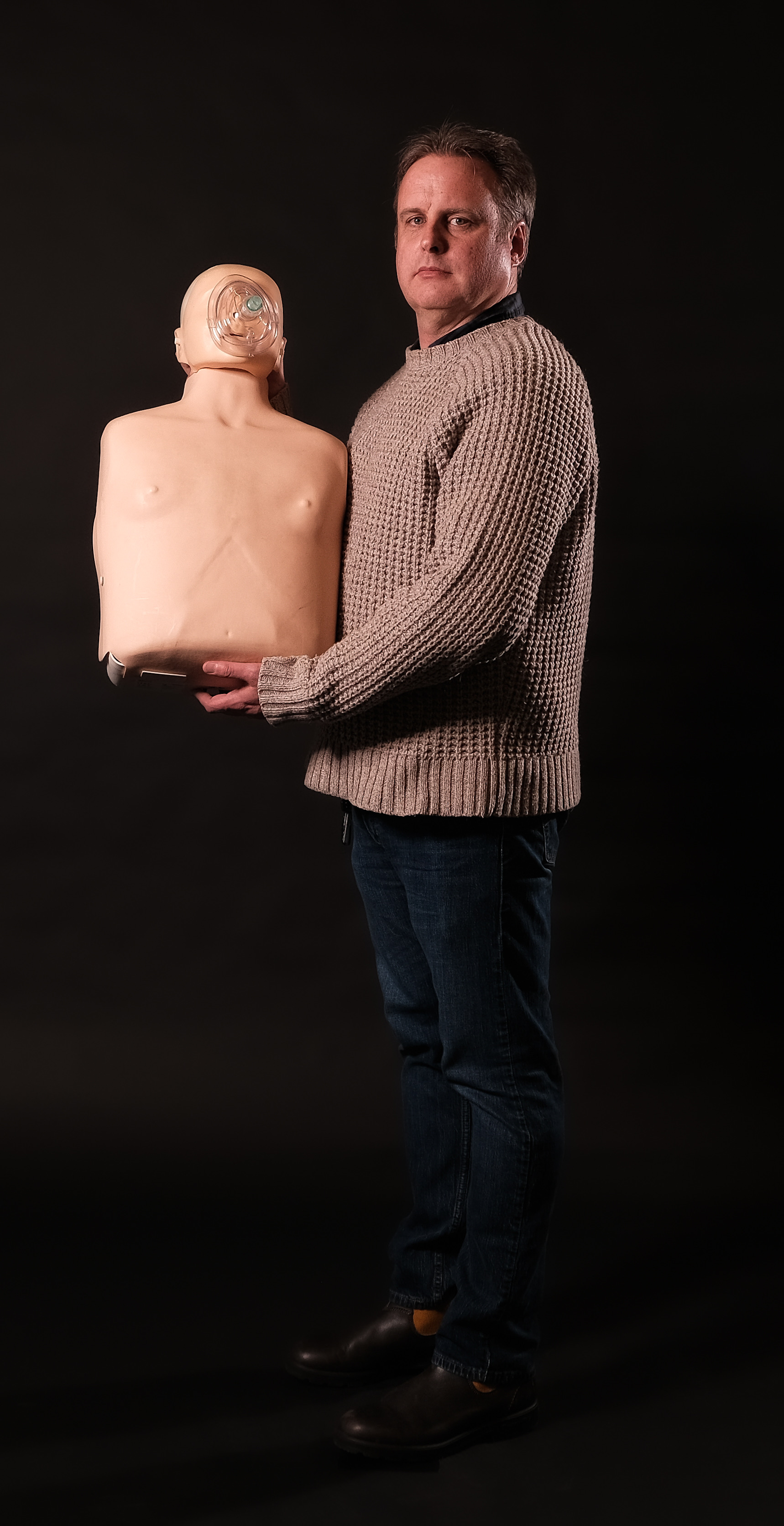Steve Willis, in a sweater and jeans, stands against a dark background holding a CPR training mannequin